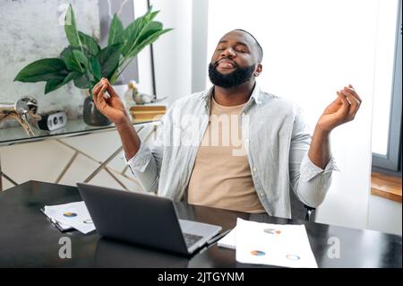 Relaxed calm peaceful African American stylish man, freelancer, designer, company ceo, taking a break, resting from online work or study in a laptop, meditating with closed eyes, relaxing, smiling Stock Photo