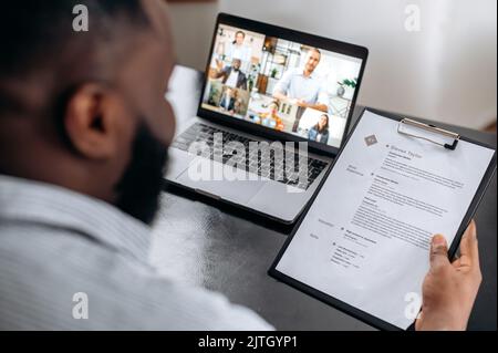 Job interview, African American company hiring manager, sits at office desk in modern office, holds resume in hand, interviews job multiracial candidates by video conference using laptop Stock Photo