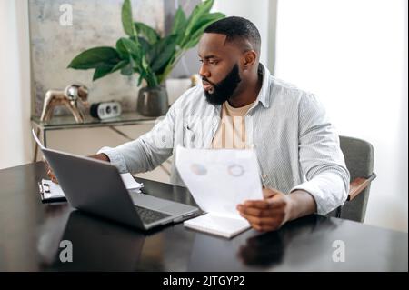 Focused busy successful african american young man, freelancer or company ceo, executive, works with documents, sit at desk in modern office, works on a project uses laptop, studying financial graphs Stock Photo