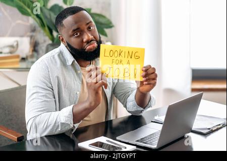 Looking for a job. Sad unemployed african american man, sit at a desk in office, lost his job, holds a sign with the inscription looking for a job, hopes to get a new job, asks for support Stock Photo