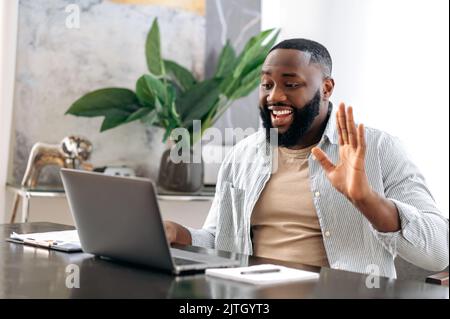 Online video conversation. Positive friendly african american man, sales manager, creative manager, sits in office, has business discussion with colleagues by video call using laptop, smiling Stock Photo