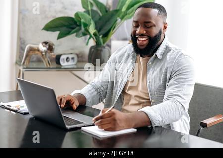 Joyful successful smart african american male company owner, sales manager, sitting at a desk in modern office, working on a project, analyzing profit, taking notes, prepares a report, smiling Stock Photo