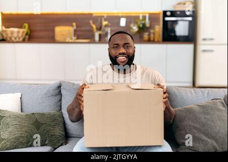 Satisfied african american man, sits on sofa in a living room, holding a large cardboard box, received a long-awaited parcel from the online store, preparing to unpack, looks at camera, smiles happily Stock Photo