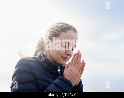 Finding strength and peace through prayer. an attractive young woman standing with her hands together in prayer outside. Stock Photo