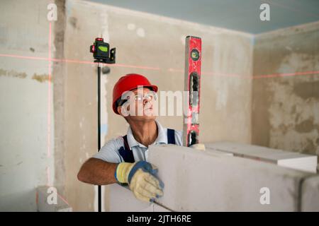 Bricklayer using spirit bubble and laser level to precise check concrete blocks on wall. Contractor uses tools for brickwork. Worker constructs a wall Stock Photo