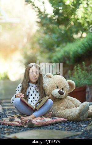 Children have the most magical imaginations. Portrait of a little girl reading a book with her teddy bear beside her. Stock Photo
