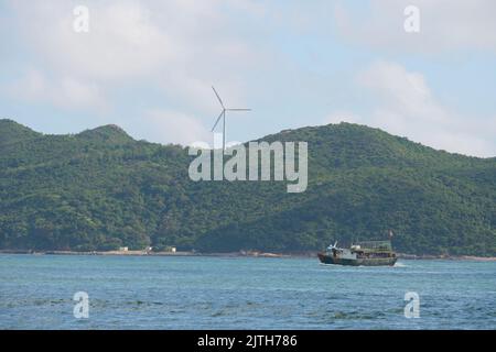 A wind turbine on an evergreen hill against a blue sky with a lake in the foreground in Lamma Island Stock Photo