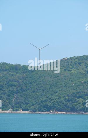 A vertical shot of a wind turbine on an evergreen hill against a blue sky in Lamma Island, Hong Kong Stock Photo