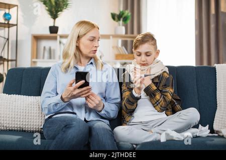 Cold and flu, virus infection. Worried mother talking with doctor by phone, while her sick teen son with scarf around neck sitting on sofa at home with a thermometer after measuring the temperature. Stock Photo