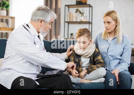 Front view of good-looking professional senior male caucasian doctor in medical coat, consulting his teen boy patient wihis mom, touching hand of chil Stock Photo