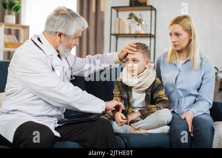 Front view of good-looking professional senior male caucasian doctor in medical coat, consulting his teen boy patient wihis mom, touching hand of child to check pulse, during home visit. Stock Photo
