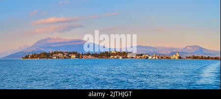 Panorama of rippled azure waters of Lake Garda with scenic skyline of Sirmione against the foggy Garda Prealps on sunset, Italy Stock Photo