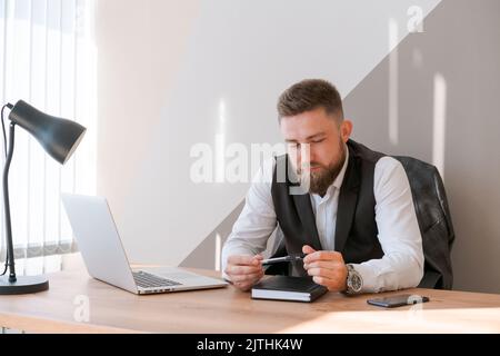 Handsome businessman is working with laptop in office, in business clothes at his desk. A bearded successful guy does his job perfectly well. Reflects and analyzes his actions Stock Photo