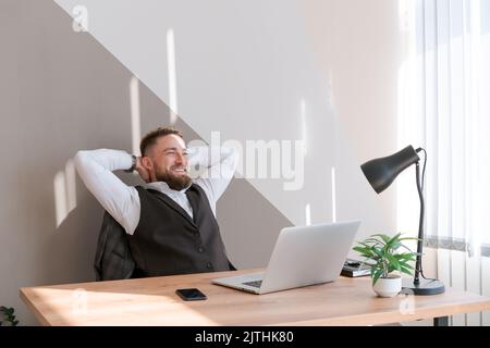 Portrait smiling man with beard, working in office on some project, he sits at table with a laptop, enjoys his success with his hands raised. Caucasian successful business guy Stock Photo