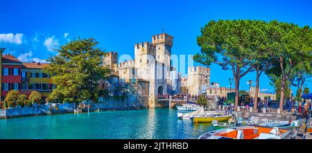 The tourist and fishing boats in a small marina of Sirmione, located next to the old town with a view on hotels, colored houses and towers of Scaliger Stock Photo