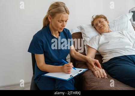 Doctor measures patient's heart rate and makes note during treatment in a hospital ward Stock Photo