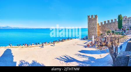 SIRMIONE, ITALY - APRIL 10, 2022: Scaligero Castle on the bank of the Lake Garda with a beach and Garda Prealps in background, on April 10 in Sirmione Stock Photo