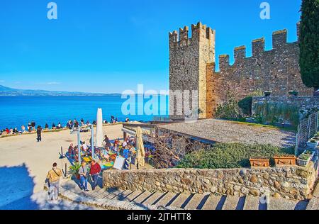 SIRMIONE, ITALY - APRIL 10, 2022: The medieval wall and tower of Scaligero Castle with Spiaggia del Prete beach and blue waters of Lake Garda, on Apri Stock Photo