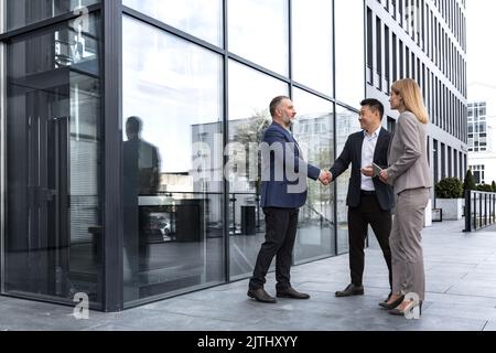 Meeting of three successful business people, diverse dream team man and woman outside office building, greeting and shaking hands, experienced professionals specialists in business suits talking Stock Photo