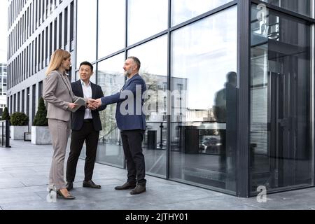 Meeting of three colleagues from outside the office building, experienced and mature IT specialists, greeting and shaking hands, business persons in business suits, diverse group of people Stock Photo