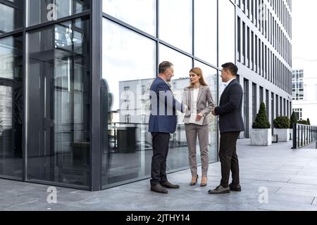 Meeting of three colleagues from outside the office building, experienced and mature IT specialists, greeting and shaking hands, business persons in business suits, diverse group of people Stock Photo