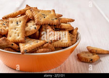 Saltine crackers in an orange bowl on a white wood table. Deep plate full of salty crackers with black and white sesame seeds close-up. Ready to eat. Stock Photo