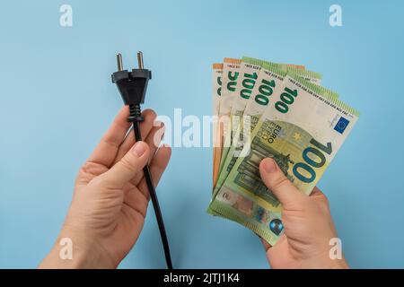 Woman hands hold electric plug and euro banknotes on blue background. High energy costs in Europe. Electric bill increases concept. Power price spike. Stock Photo