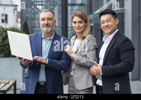 Portrait dream team IT specialists professionals team leaders outside office building looking at camera holding laptop discussing successful application project, specialists business suits smiling. Stock Photo