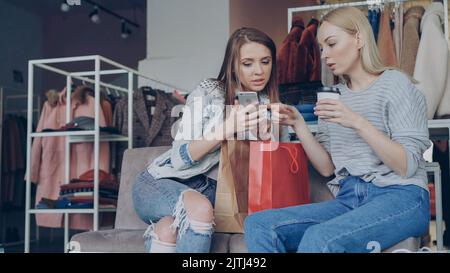 Attractive young ladies sitting on chairs in a clothing store with coffee and shopping bags, checking smartphone and talking. Shelves and hangers with colourful clothes in background Stock Photo