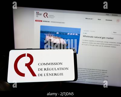 Person holding cellphone with logo of French Commission de Regulation de l'Energie (CRE) on screen in front of webpage. Focus on phone display. Stock Photo