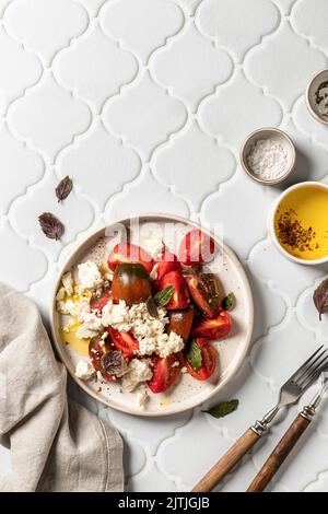 Tomato salad with feta cheese and bazil leaves served with cutlery and linen napkin on white tile background top view with text space. Flat lay, verti Stock Photo