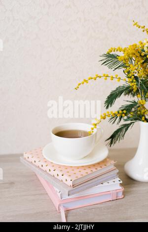 Bouquet of mimosa in a white vase, a cup of tea on a stack of notebooks for notes Stock Photo