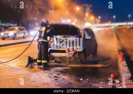 Group of fire men extinguishing and put out burning car crash after road traffic incident, fire fighting operation in the night city, firefighters wit Stock Photo