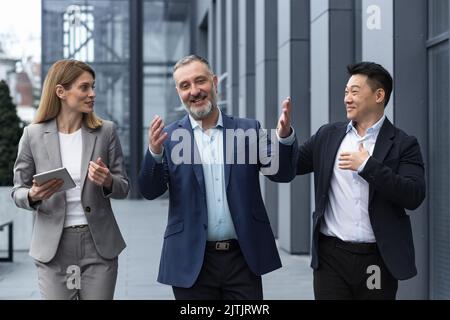 A diverse team of IT specialists, senior and experienced engineers managers team leaders, a group of three workers happily strolling outside an office building, colleagues in business suits. Stock Photo