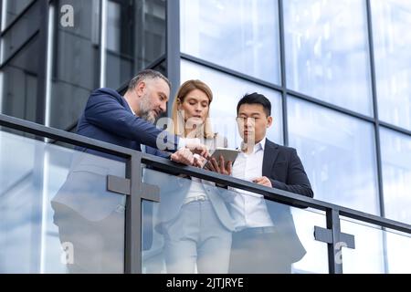 Specialist IT team leaders, group of business people outside office building, discussing and talking, testing new app on tablet, professional colleagues in business clothes, Stock Photo