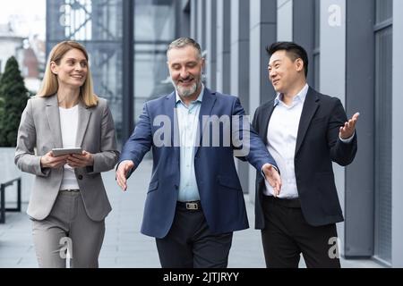 A diverse team of IT specialists, senior and experienced engineers managers team leaders, a group of three workers happily strolling outside an office building, colleagues in business suits. Stock Photo