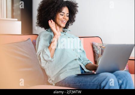 Video conference. Smiling young african american woman using laptop for making video call at home, waving at webcamera, sitting on the sofa Stock Photo