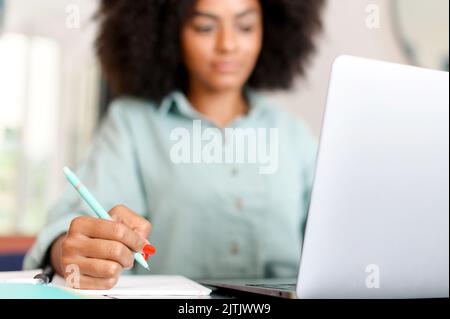 Female student or employee using laptop and taking notes in notebook. Selective focus at the pen, face is out of focus Stock Photo
