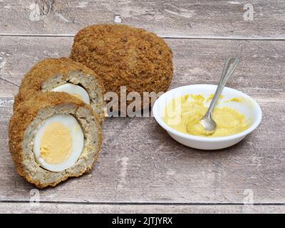 Scotch Eggs halved on a wooden table with English Mustard in a white bowl and spoon Stock Photo