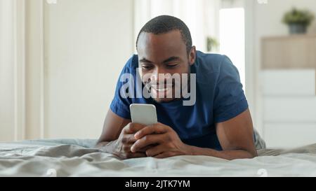 African Guy Texting On Smartphone Lying On Bed In Bedroom Stock Photo