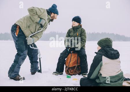 Happy mature man using ice auger while talking with son sitting on backpack at frozen lake Stock Photo