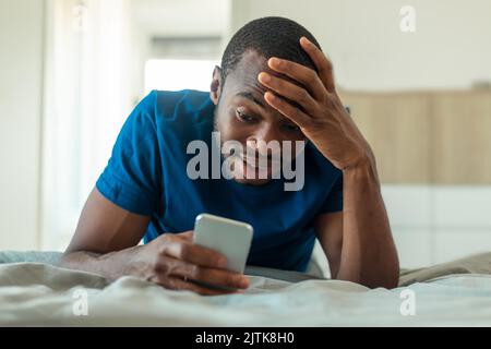 Shocked Black Man Texting On Phone Lying In Bedroom Stock Photo