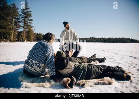 Father and son sitting on animal skin while ice fishing with friends enjoying sunny day in winter Stock Photo