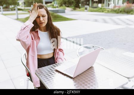 Young woman with laptop with facepalm gesture, feeling regret, sorrow, blaming herself for mistake, raise her hand, sitting in street cafe. Freelance, online business, online work. Copy space Stock Photo