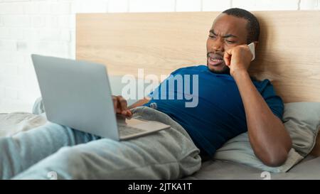 Discontented Black Guy Talking On Cellphone Using Laptop At Home Stock Photo
