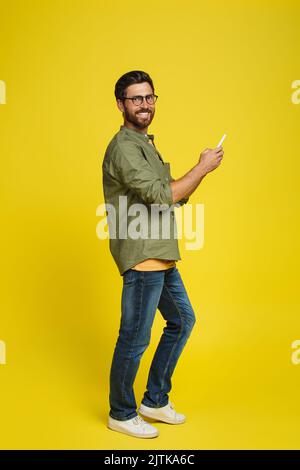 Positive caucasian man using smartphone and smiling at camera on yellow studio background, full length shot Stock Photo