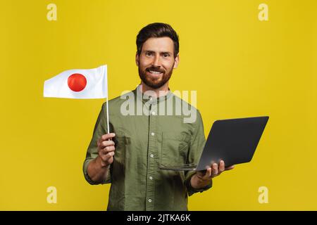 Happy man holding Japan flag and laptop computer, standing over yellow studio background Stock Photo