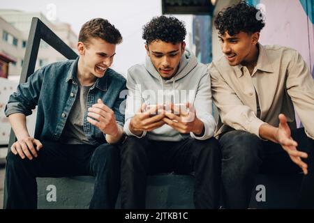 Young men looking at friend using smart phone while sitting outdoors Stock Photo