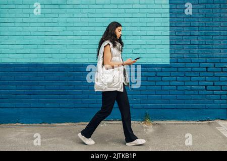 Young woman using mobile phone walking by blue brick wall Stock Photo