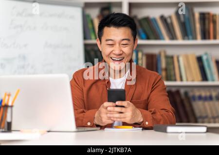 Japanese Teacher Man Using Cellphone Sitting At Desk In Classroom Stock Photo